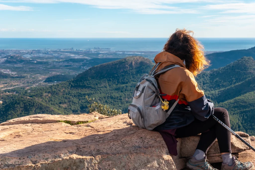 Ei dame i turklær sitter på fjellet over en tett skog og nyter utsykten med sekk på ryggen.